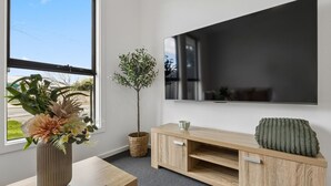 A modern living room with a flat-screen TV mounted on the wall, a wooden entertainment unit with a cushion, a potted plant, and a vase with flowers on a wooden table. - Modern four bedroom home with everything you could possibly need in a prime coast to city location! (South Plympton)