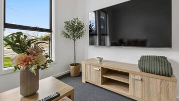 A modern living room with a flat-screen TV mounted on the wall, a wooden entertainment unit with a cushion, a potted plant, and a vase with flowers on a wooden table.