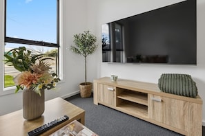 A modern living room with a flat-screen TV mounted on the wall, a wooden entertainment unit with a cushion, a potted plant, and a vase with flowers on a wooden table. - Modern four bedroom home with everything you could possibly need in a prime coast to city location! (South Plympton)