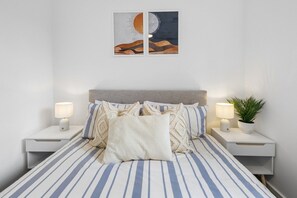 Ein ordentlich bezogenes Bett mit gestreifter Bettwäsche, zwei Nachttische mit Lampen und ein an der Wand hängendes Kunstwerk, das Sonne und Mond zeigt.
