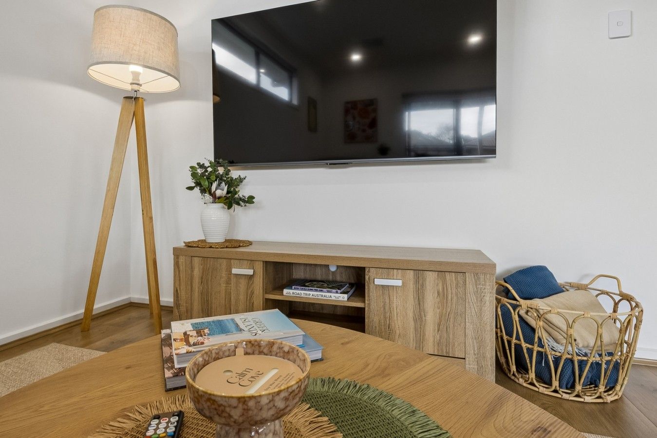 A modern living room with a flat-screen TV mounted on the wall, a wooden entertainment unit with books, a floor lamp, a wicker chair, and a decorative bowl on a wooden table.