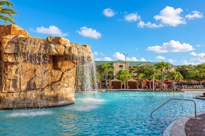A pool with a waterfall feature, surrounded by lounge areas and a building with a green roof.