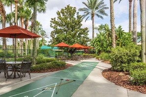 A croquet court with a green surface, surrounded by palm trees and outdoor seating with red umbrellas.