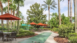 A croquet court with a green surface, surrounded by palm trees and outdoor seating with red umbrellas.