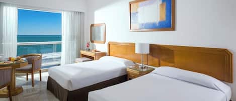 A hotel room with two beds, a desk with a chair, a bedside table, a lamp, and a large window offering a view of the ocean.