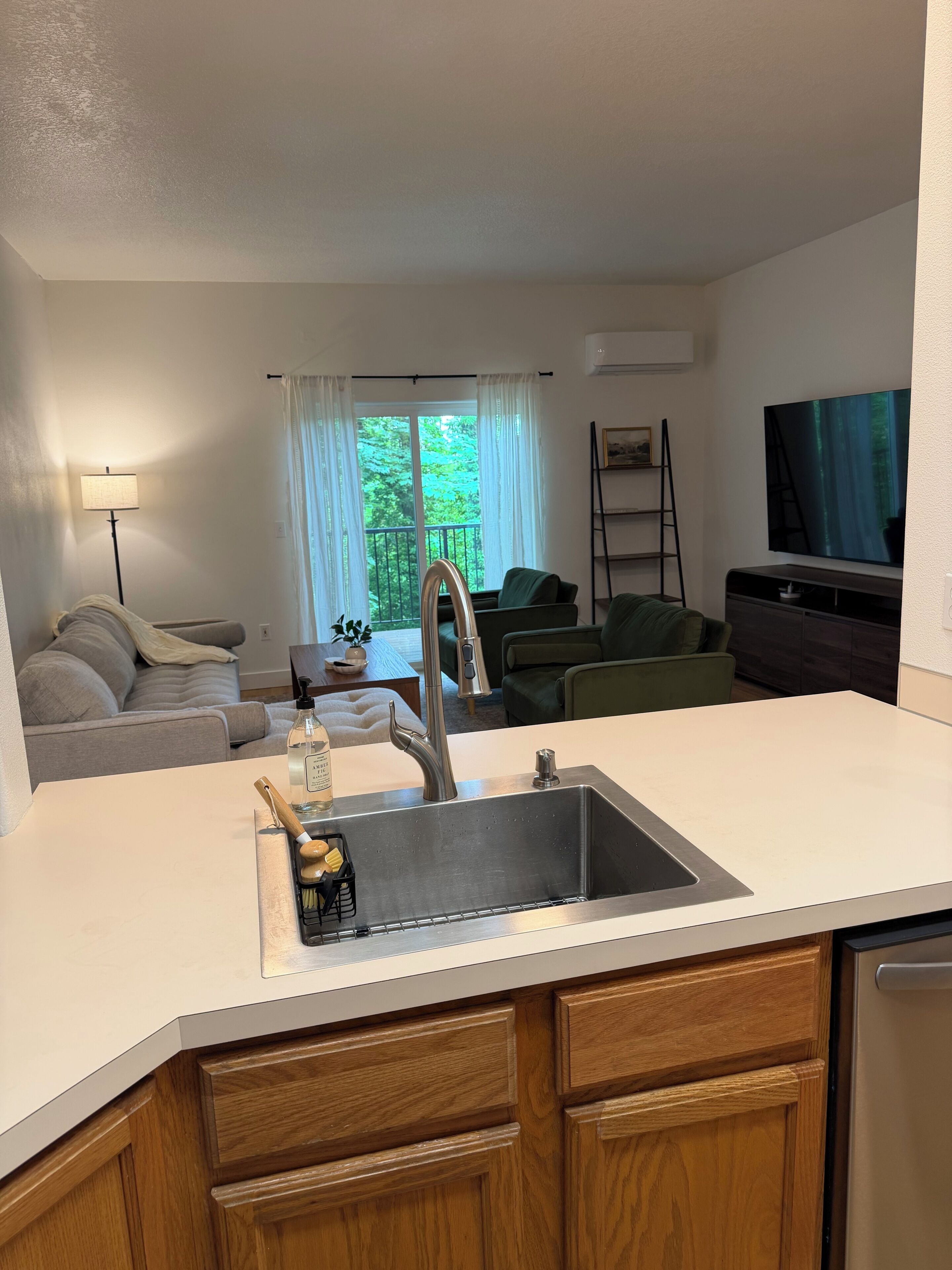 A modern kitchen with a stainless steel sink, wooden cabinets, a dining area with a table and chairs, a sofa, and a television.