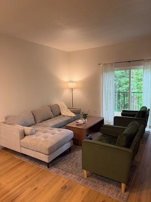 Uma sala de estar moderna com sofá modular, uma poltrona verde, mesa de centro e abajur. Há uma janela grande com cortinas e vista para área verde.