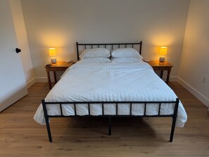 Uma cama com colcha branca, duas mesas de cabeceira com luminárias e piso de madeira.