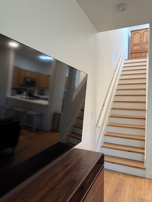 Interior moderno com escada de madeira, televisão de tela plana em suporte de madeira e área de cozinha ao fundo.
