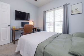 A bedroom with a bed, a desk with a computer, a television on the wall, and a window with curtains.