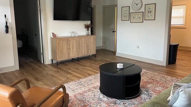 Een moderne woonkamer met een flatscreen televisie, een houten kast, een ronde zwarte salontafel en een kamerkleed met dessin.