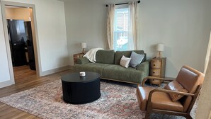 Een woonkamer met een bank, een salontafel, een stoel, een kamerkleed en een raam met gordijnen.