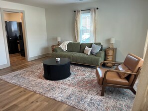 Una sala de estar con sofá, mesa de centro, una silla, alfombra estampada y ventana con cortinas.
