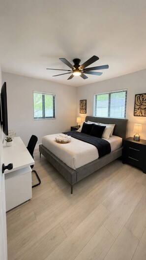 Quarto moderno com cama, mesas de cabeceira, escrivaninha e ventoinha no teto.