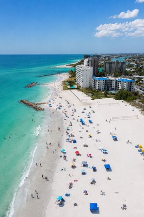 Uma praia com vários guarda-sóis e pessoas, céu azul límpido e edifícios ao fundo.