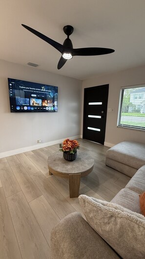 A living room with a flat-screen TV, a ceiling fan, a coffee table with a flower arrangement, and a sofa. - The Lincoln Luxe • Midtown Tampa •Near Downtown • Raymond James Stadium• Airport (Tampa)