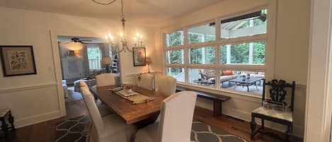 A dining area with a wooden table, white chairs, and a chandelier. There is a large window with a view of greenery outside.