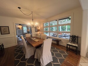 A dining area with a wooden table, white chairs, and a chandelier. There is a large window with a view of greenery outside.