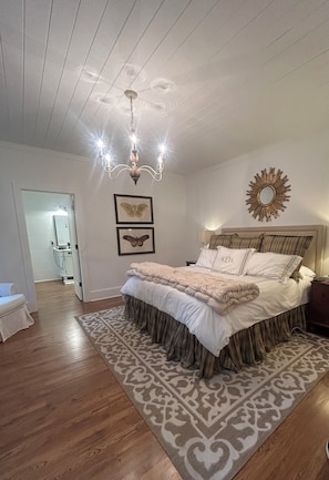 A bedroom with a large bed, a chandelier, a patterned rug, and a mirror on the wall.
