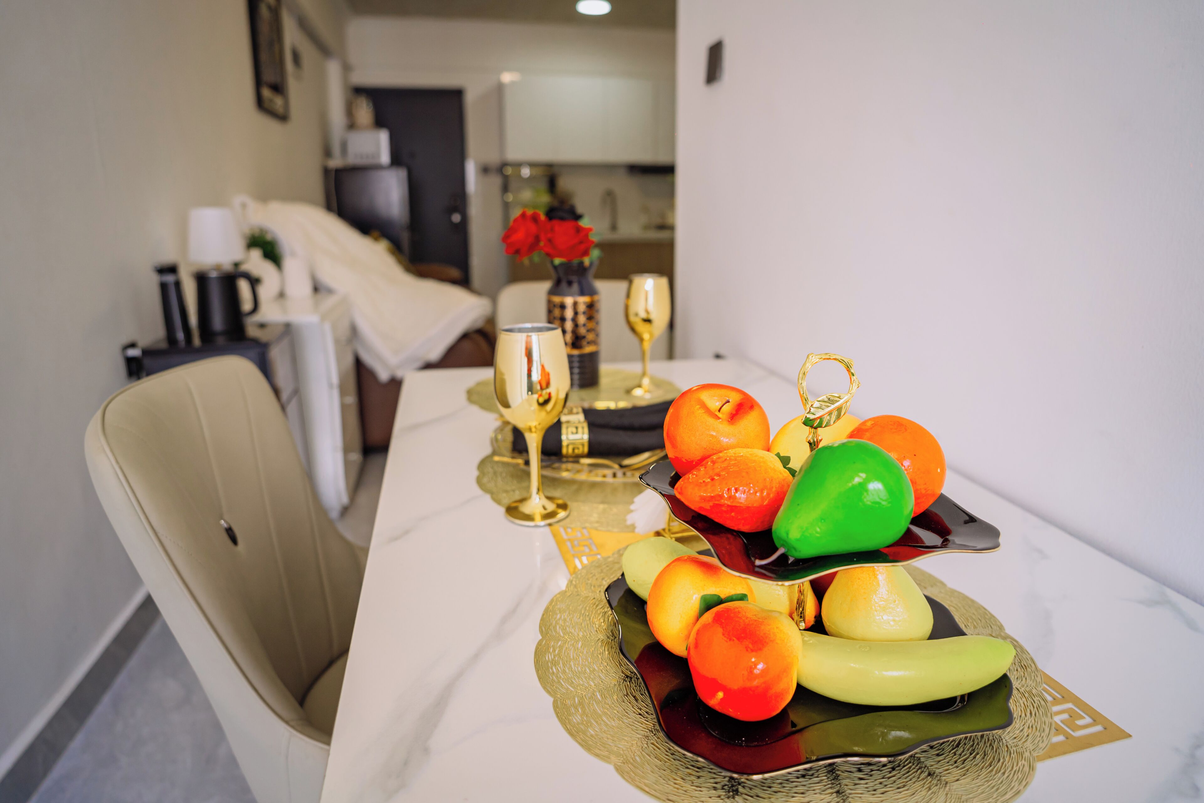 A dining table with a fruit platter, two wine glasses, and a vase with red roses.