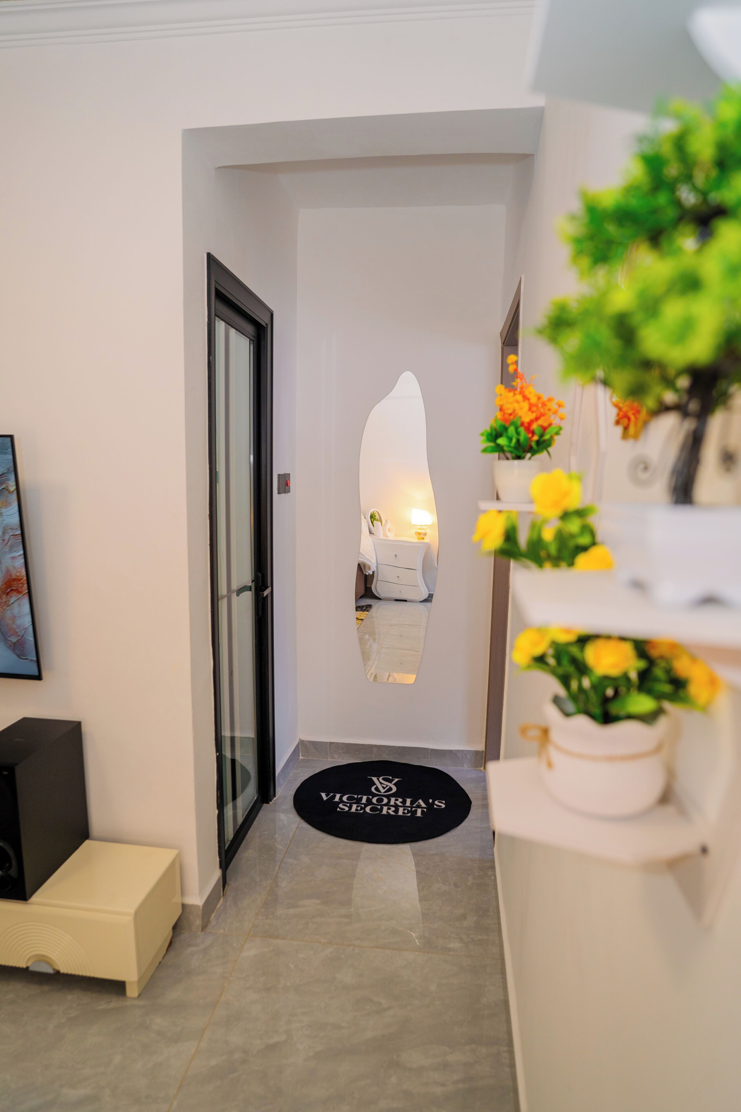 Um corredor com porta de vidro, um tapete preto com texto, um armário branco com flores amarelas e um quadro na parede.