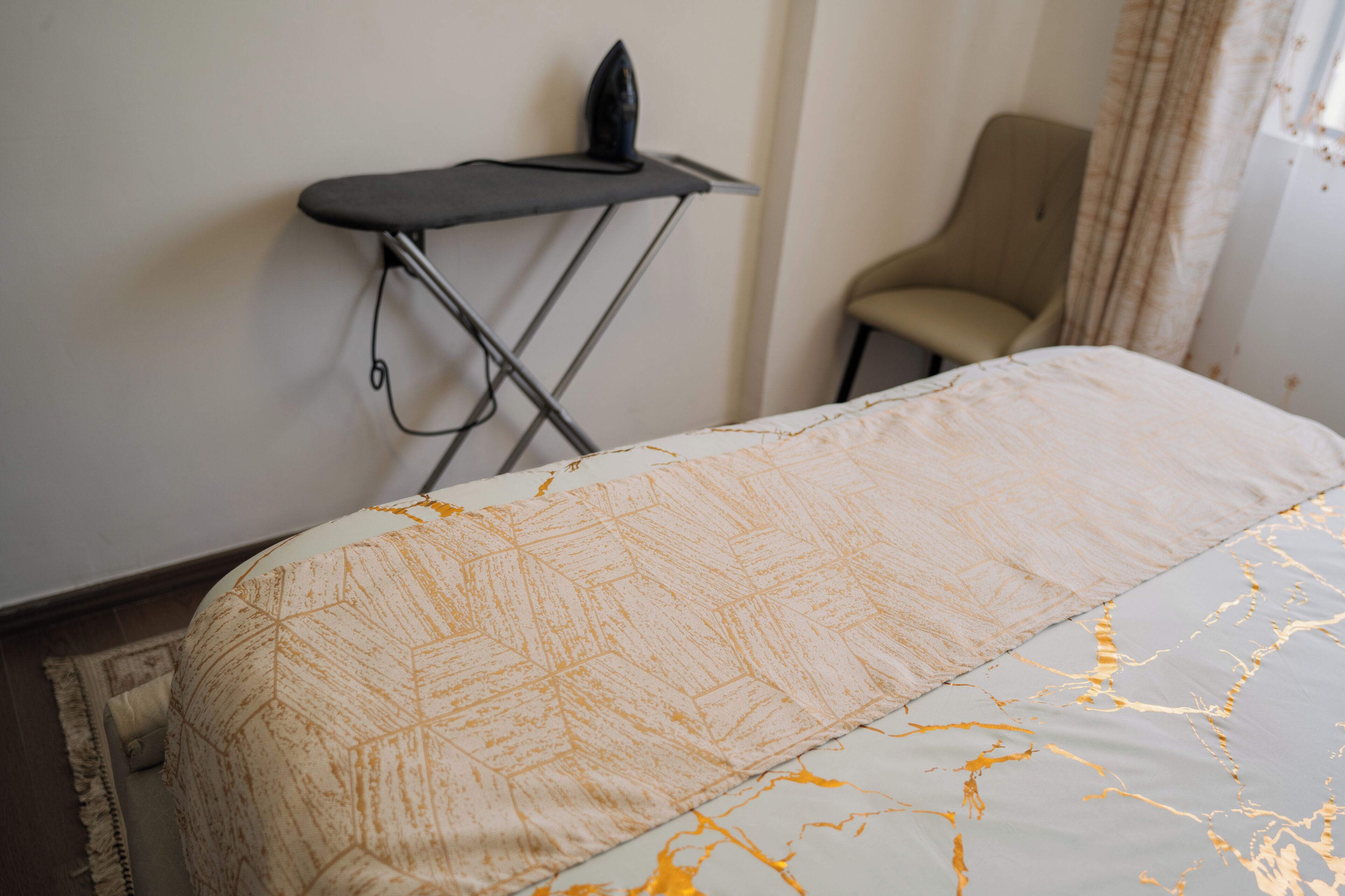 Un lit bien fait, recouvert d’une couvre-lit à motifs, un petit fer à repasser et une chaise adossée au mur.