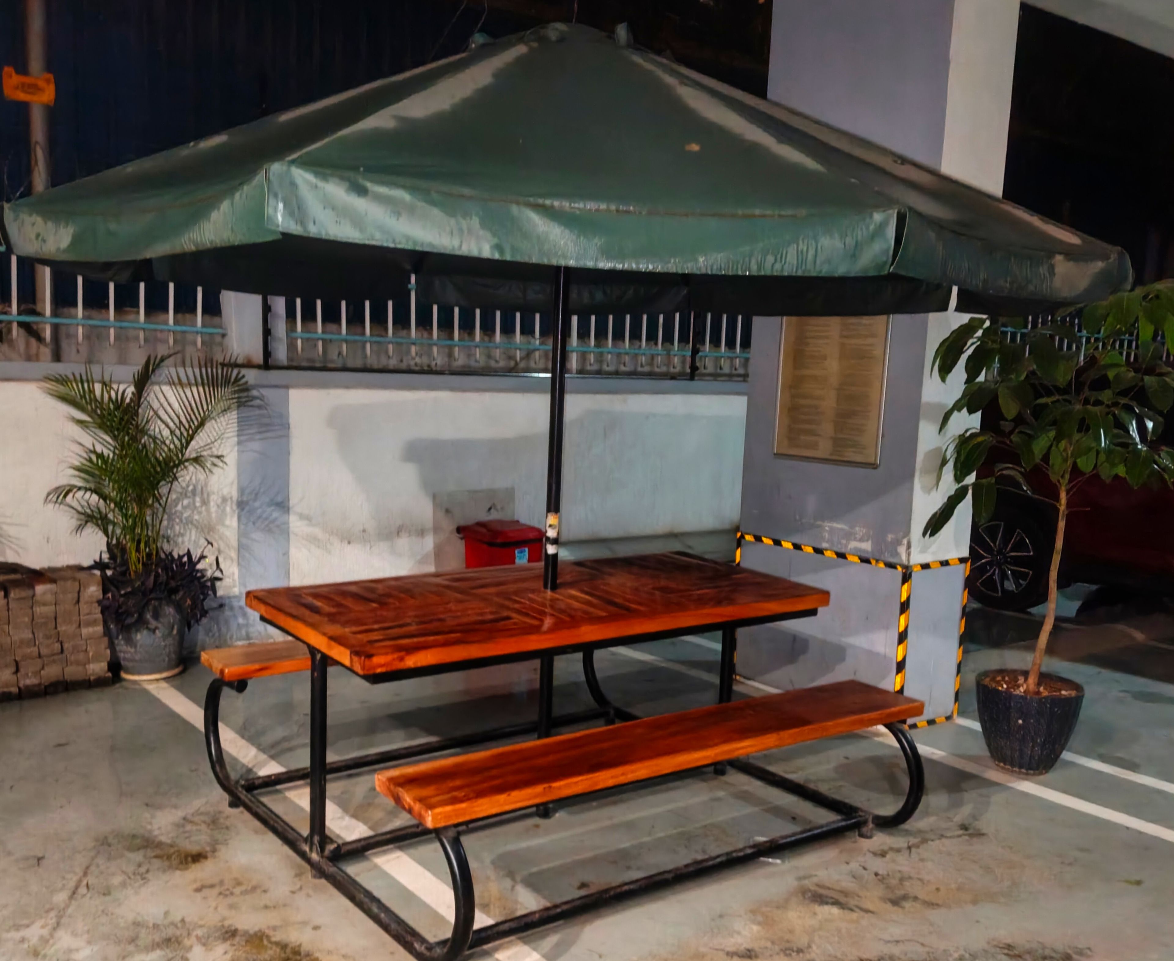 A wooden picnic table with a green umbrella, surrounded by potted plants and a metal fence.