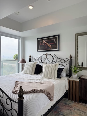 A bedroom with a large bed, a headboard with a decorative design, a framed picture on the wall, a bedside table with a lamp, and a view through the window.