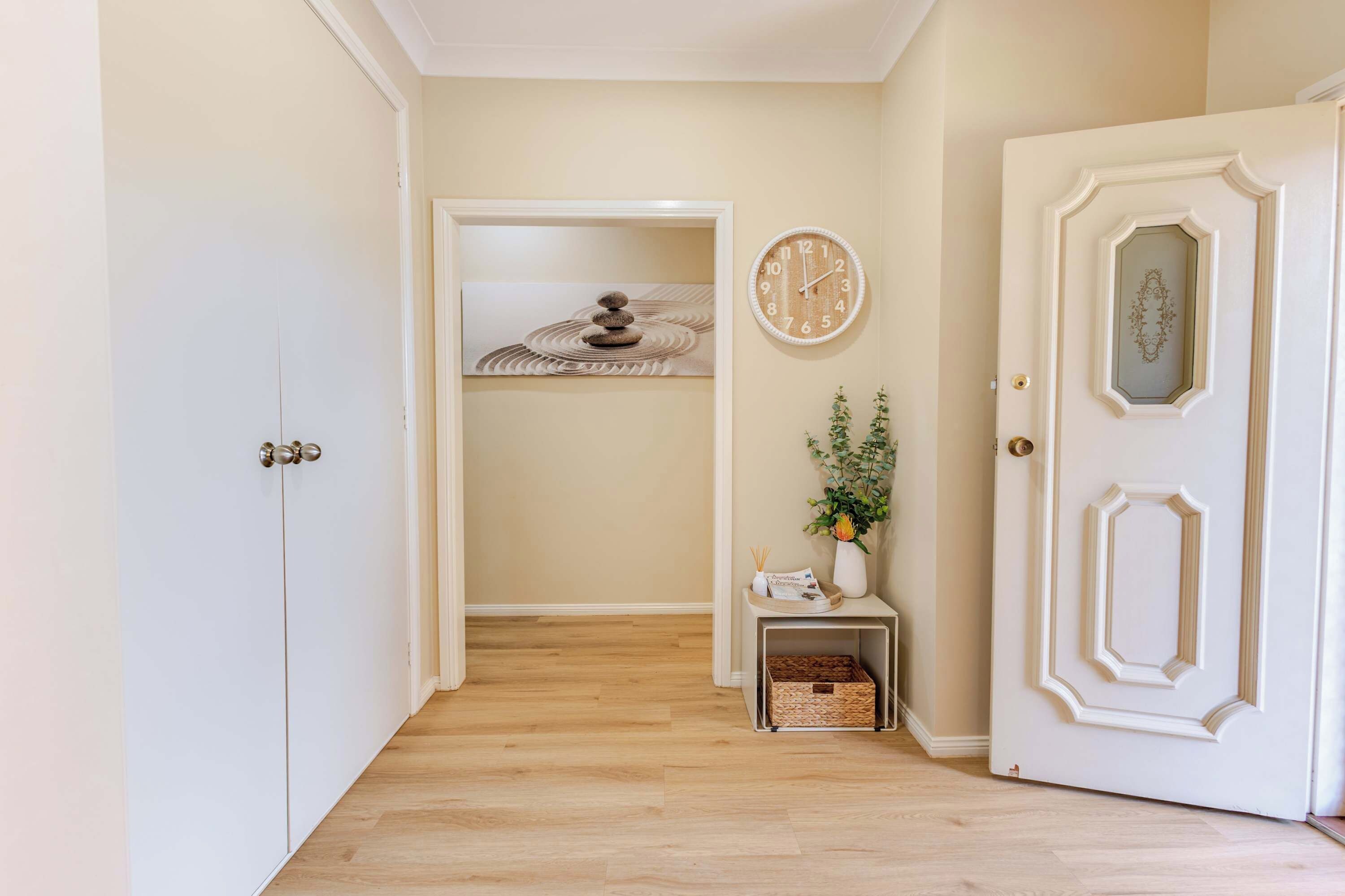 A hallway with a wooden floor, a white door with a decorative panel, a wall clock, and a small table with a vase and a basket.