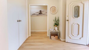 A hallway with a wooden floor, a white door with a decorative panel, a wall clock, and a small table with a vase and a basket. - House of Tudor II, Dubbo (Dubbo)