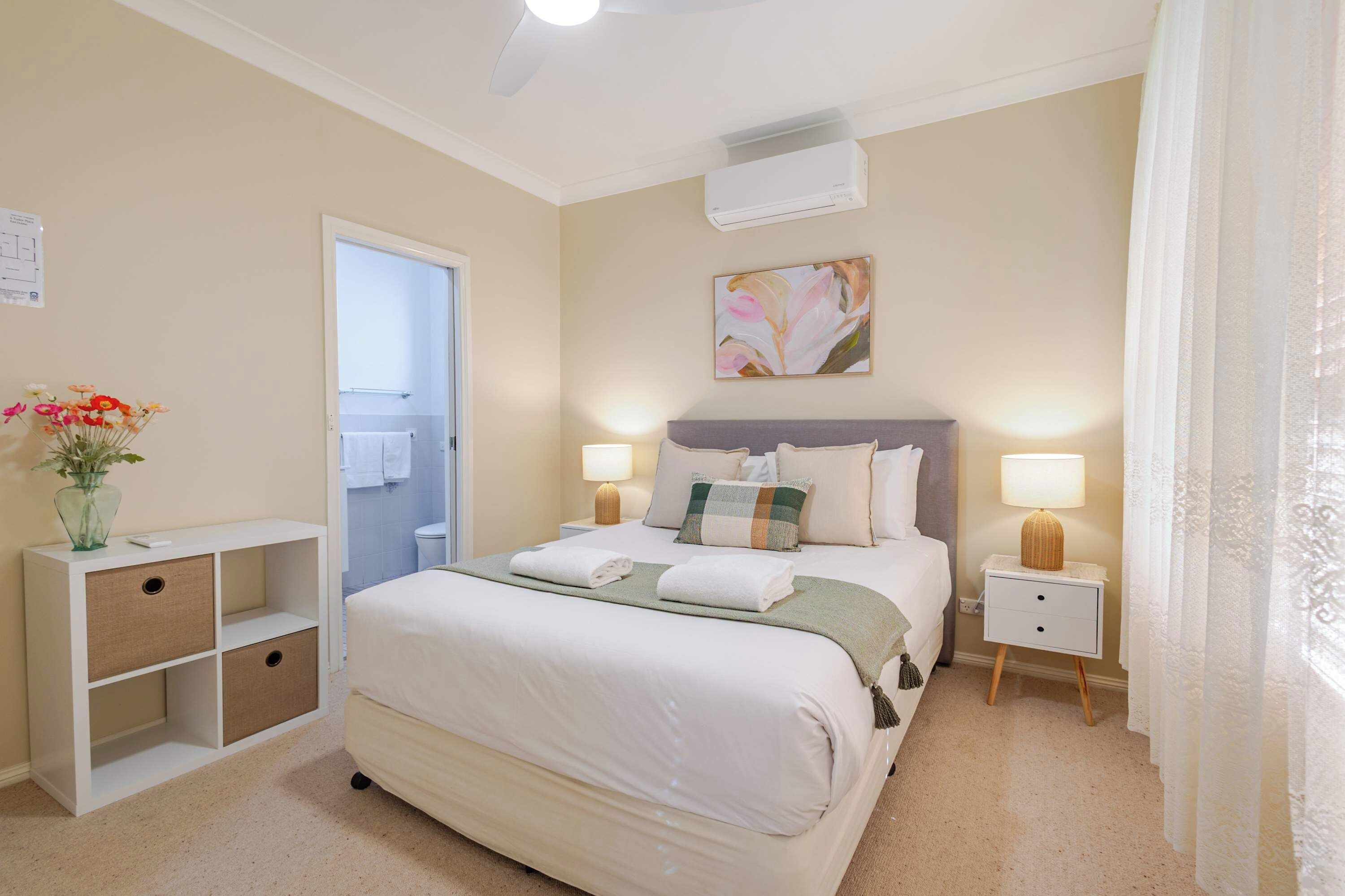 A hotel room with a bed, bedside tables, a nightstand, a wall-mounted air conditioner, and a bathroom visible through an open door.