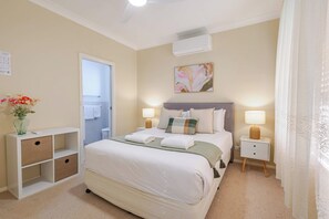 A hotel room with a bed, bedside tables, a nightstand, a wall-mounted air conditioner, and a bathroom visible through an open door. - House of Tudor II, Dubbo (Dubbo)