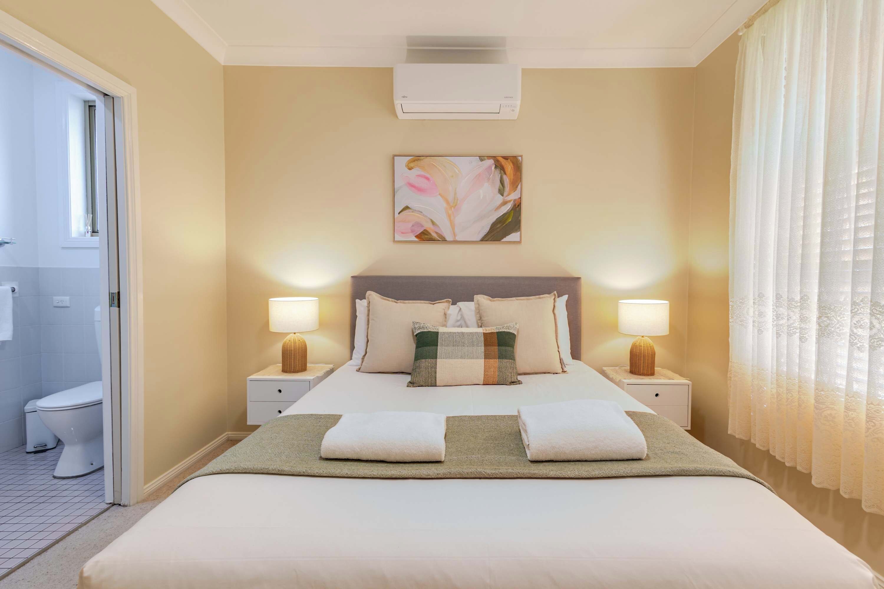 A hotel room with a bed, two bedside tables with lamps, a wall-mounted air conditioner, and a window with curtains.