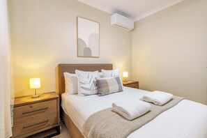 A hotel room with a bed, two bedside tables, a lamp, a wall-mounted air conditioner, and a framed picture. - House of Tudor II, Dubbo (Dubbo)