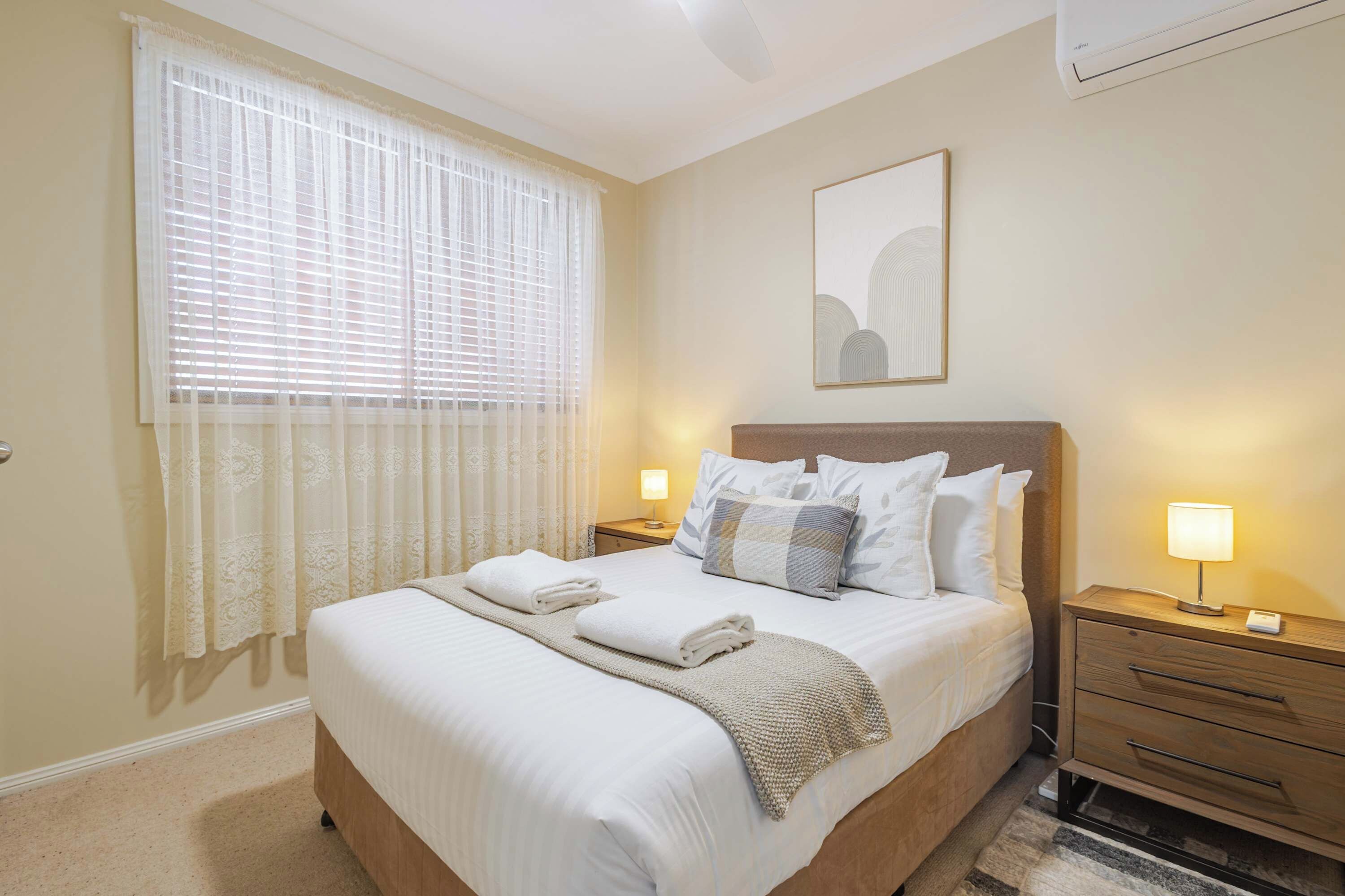 A hotel room with a bed, two bedside tables with lamps, a window with sheer curtains, a wall-mounted air conditioner, and a framed artwork above the bed.