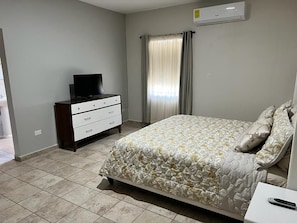 Une chambre à coucher avec un lit, un téléviseur posé sur une commode, une fenêtre avec des rideaux et un climatiseur fixé au mur.
