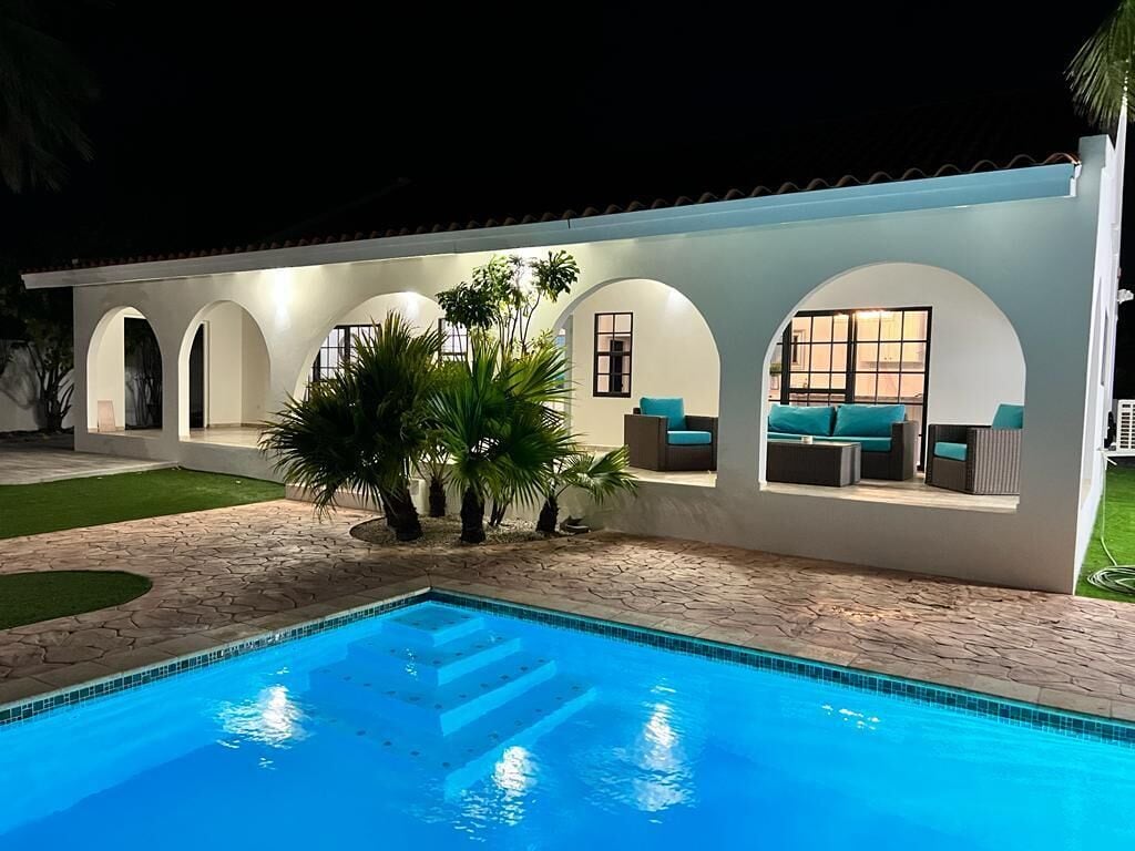 A swimming pool with a tiled edge, a covered outdoor seating area with cushions, and a building with arched windows.