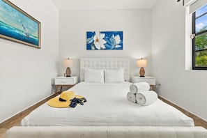 Une chambre avec un lit blanc, deux tables de chevet avec des lampes, un tableau au-dessus du lit et une fenêtre sur le côté droit.