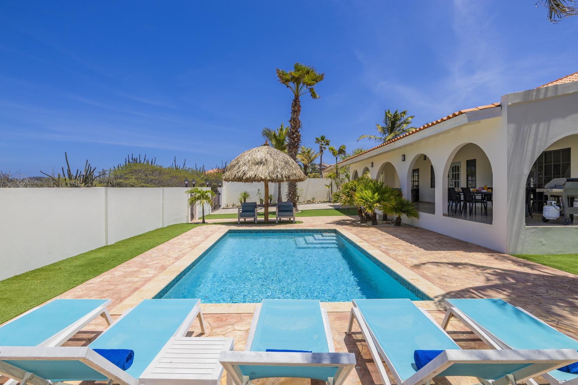 A swimming pool with lounge chairs, a thatched-roof structure, and a dining area with outdoor seating.
