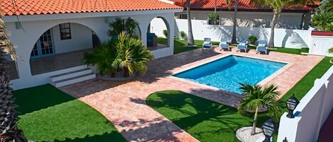 Une piscine avec une terrasse carrelée, entourée de palmiers et d’un bâtiment blanc doté d’un toit rouge.