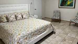 A bedroom with a large bed, a nightstand, a chair, a door, and a painting on the wall. - Renovated 3BR Home Private PoolClose 2 BeachBBQ (Noord)