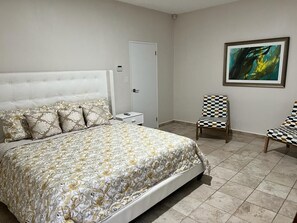 Ein Schlafzimmer mit einem großen Bett, einem Nachttisch, einem Sessel, einer Tür und einem Bild an der Wand.