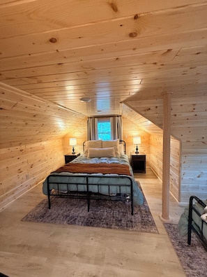 A cozy attic bedroom with a bed centered in the room, flanked by nightstands with lamps, and a window at the far end.