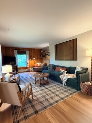 Una sala de estar acogedora con sofá, sillón, mesa de centro, pisos de madera y un ventanal grande.