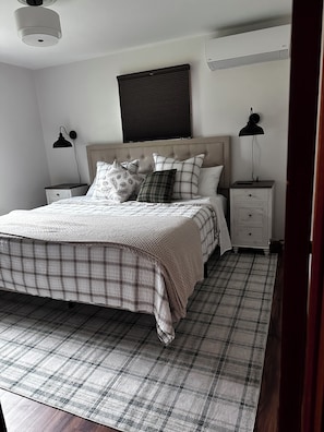 A bedroom with a bed, two bedside tables, a headboard, a wall-mounted air conditioner, and a ceiling light fixture.