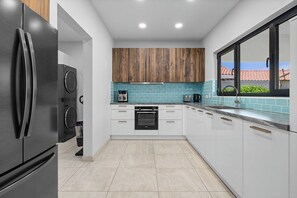 A modern kitchen with white cabinets, a black refrigerator, a built-in oven, and a window with a view of a red-tiled roof. - Villa Prikichi II ~ Close to Beach w/ Private Pool (Noord)