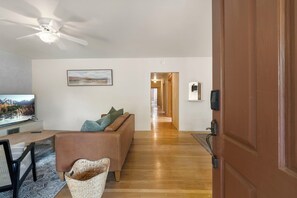 A living room with a sofa, a television, a ceiling fan, and a hallway leading to another room. - Entire Home-Charming Historic Adobe Prime Location (Santa Fe)