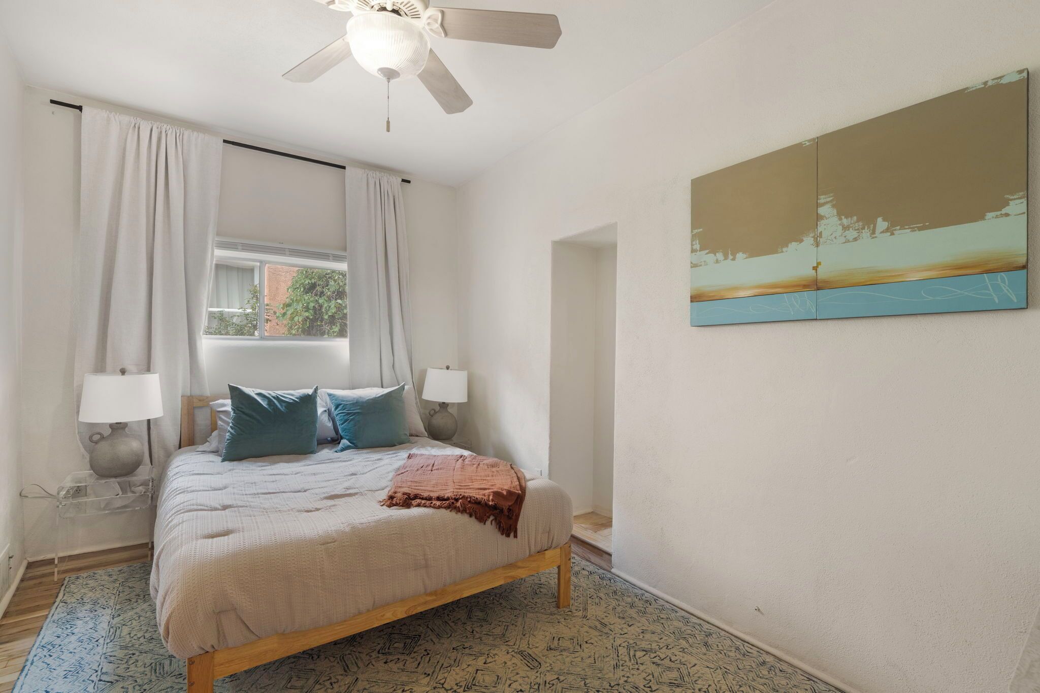 Un dormitorio con cama, dos lámparas de noche, ventilador de techo, ventana con cortinas y un cuadro en la pared.