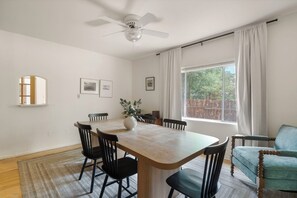 A dining area with a wooden table, black chairs, a ceiling fan, and a view of a wooden fence through a window. - Entire Home-Charming Historic Adobe Prime Location (Santa Fe)