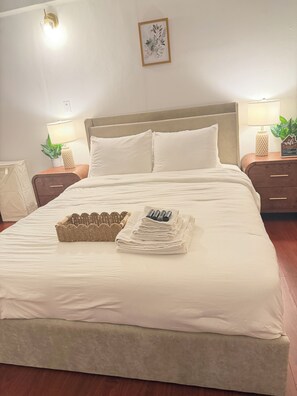 A neatly made bed with white linens, a woven basket, and folded towels on a room with wooden flooring. - House of VANTA on Fifteenth (Miami)